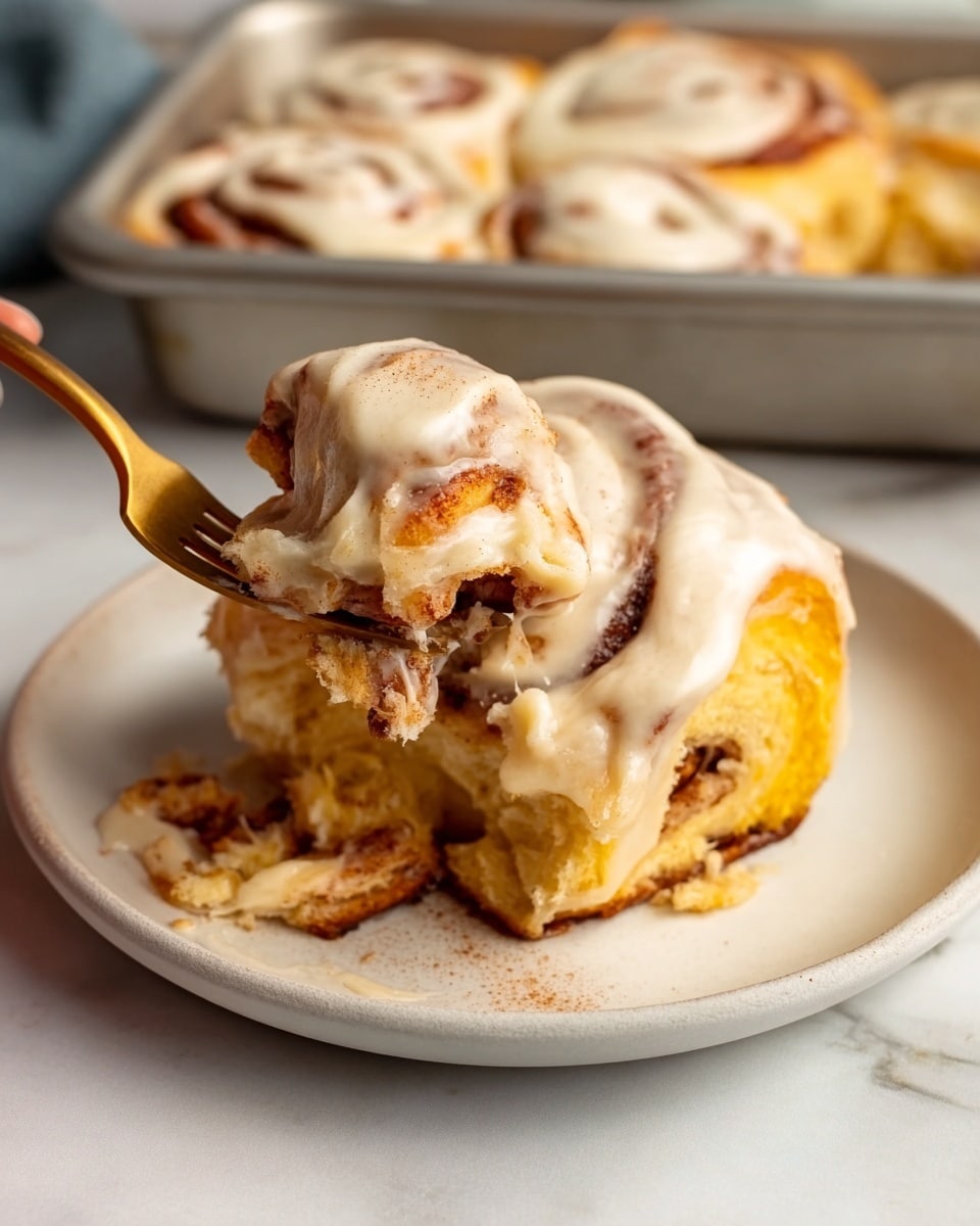 A close-up of a three-layer cinnamon roll with bright yellow dough, dark brown swirls of cinnamon filling, and a thick, creamy off-white frosting spread over the top. The cinnamon roll is sitting on a small white plate, with a portion lifted by a gold spoon showing the soft, fluffy interior and gooey filling inside. Some cinnamon powder is lightly dusted on the frosting and plate edge. In the background, a white tray holds more cinnamon rolls. The surface beneath is a white marbled texture. photo taken with an iphone --ar 4:5 --v 7