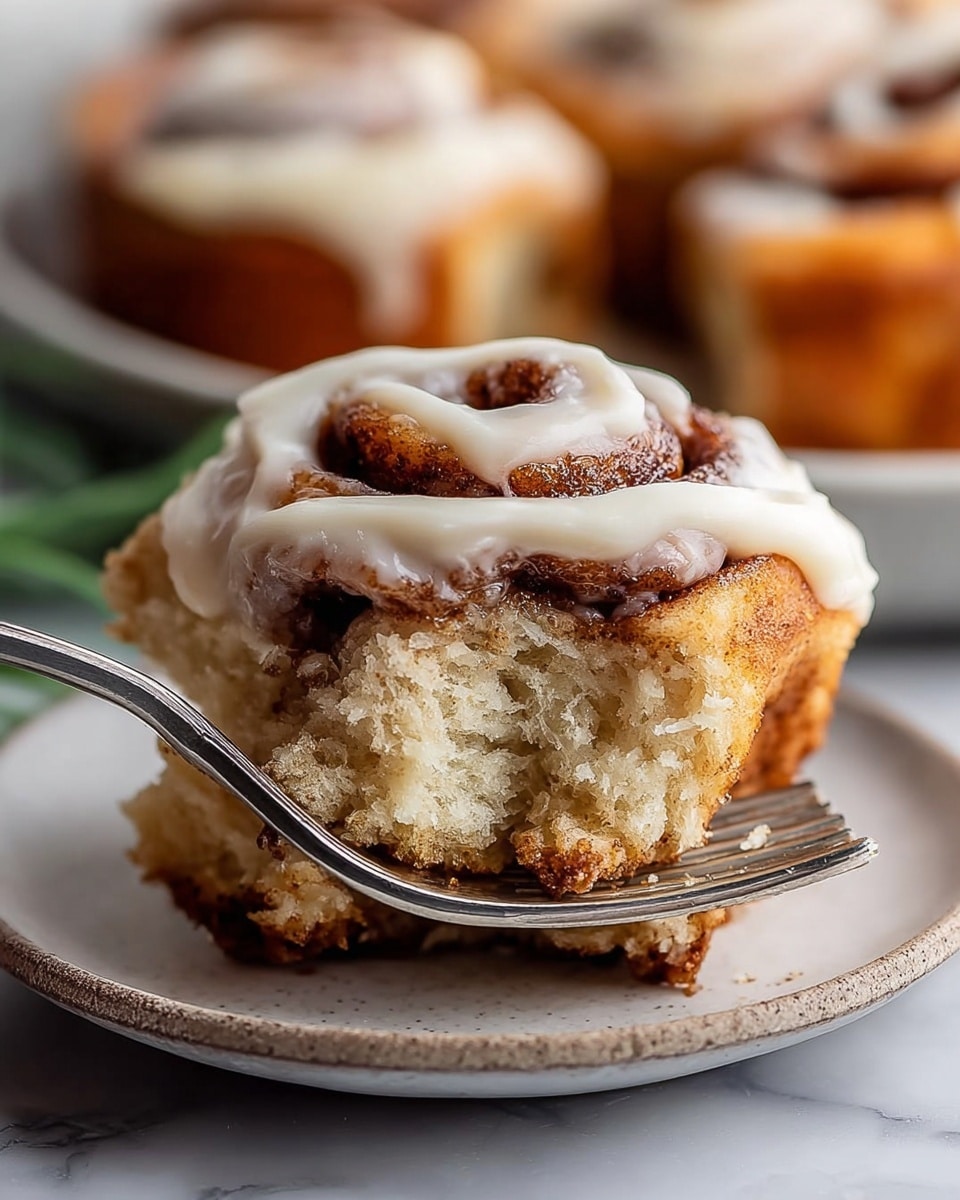 A close-up view of a single cinnamon roll held by a silver fork, showing three visible layers: a golden-brown crust layer at the bottom with a slightly crispy texture, a soft, light beige dough layer in the middle with a slightly crumbly look, and a top spiral layer with dark brown cinnamon swirls covered partially by a creamy white icing that is smooth and thick, placed on a white plate with a slightly rough edge, all set on a white marbled surface with blurred cinnamon rolls in the background, photo taken with an iphone --ar 4:5 --v 7