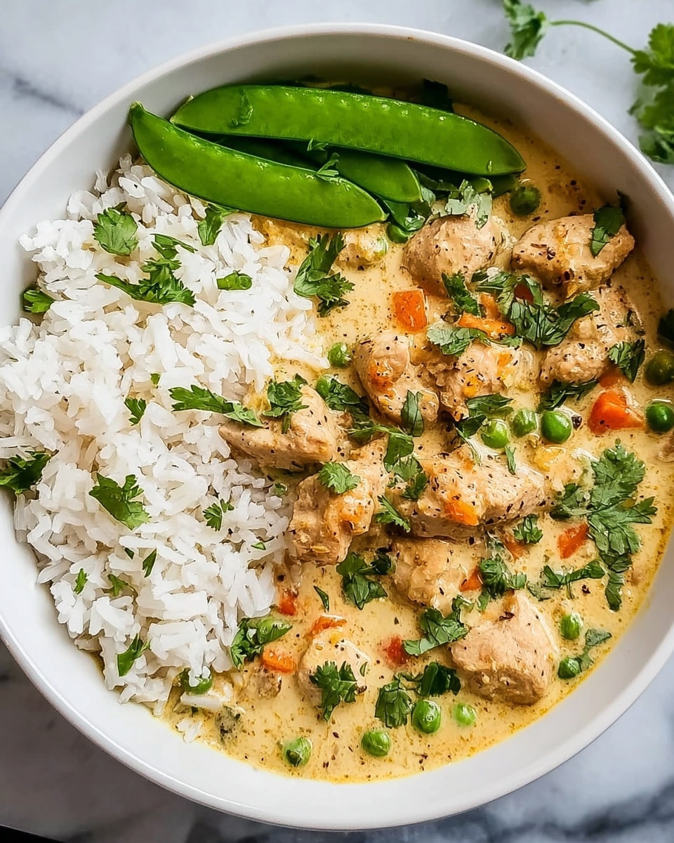 The image shows a white bowl filled with three distinct layers. On the left side, there is a layer of plain white rice with visible grains. The top right half is covered with a creamy, light yellow curry sauce with chunks of browned chicken pieces scattered all over. The curry also has small green peas and bits of orange carrot mixed in, and fresh green cilantro leaves are sprinkled on top. Along the top edge of the bowl, there is a small portion of bright green snap peas. The bowl is placed on a white marbled surface. photo taken with an iphone --ar 4:5 --v 7
