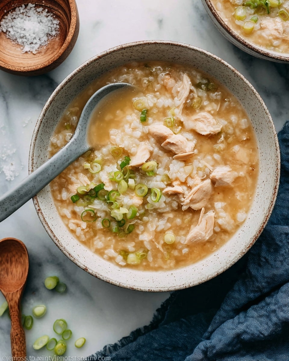A white speckled bowl filled with a thick, golden-brown rice soup with soft, light beige pieces of chicken mixed throughout. Bright green chopped scallions are sprinkled on top, adding small pops of color across the surface. A gray ceramic soup spoon rests inside the bowl on the left side, partially submerged in the soup. The bowl sits on a white marbled surface with another similar bowl partly visible in the background. photo taken with an iphone --ar 4:5 --v 7