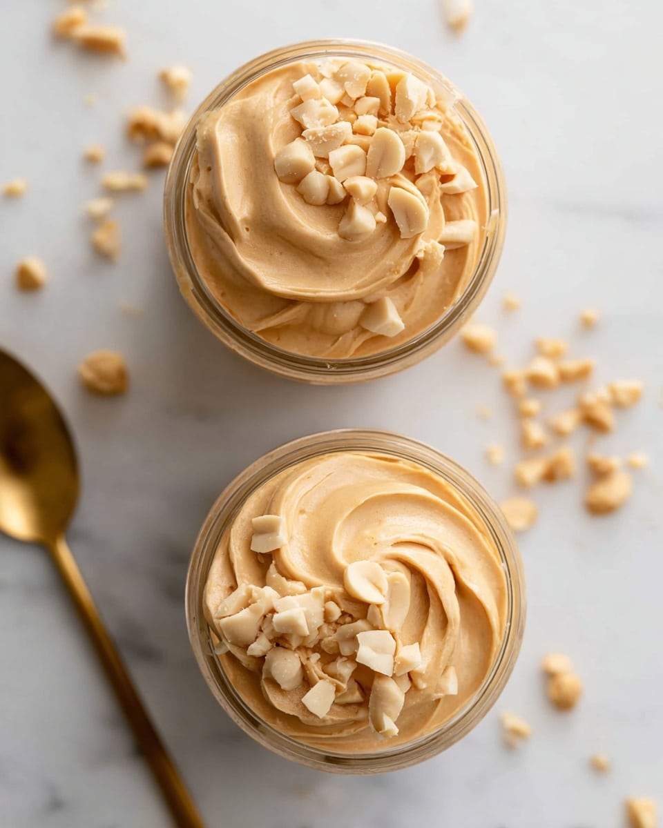 Two clear jars sit on a white marbled surface, each filled with smooth, light brown peanut butter cream swirled into soft peaks. The peanut butter is topped with chunky pieces of pale beige peanuts, scattered unevenly on one side of the swirl. Small peanut bits are also scattered around the jars on the surface. A gold spoon is partially visible near the bottom left corner of the image. The overall look is creamy and rich with a natural nutty texture. Photo taken with an iphone --ar 4:5 --v 7