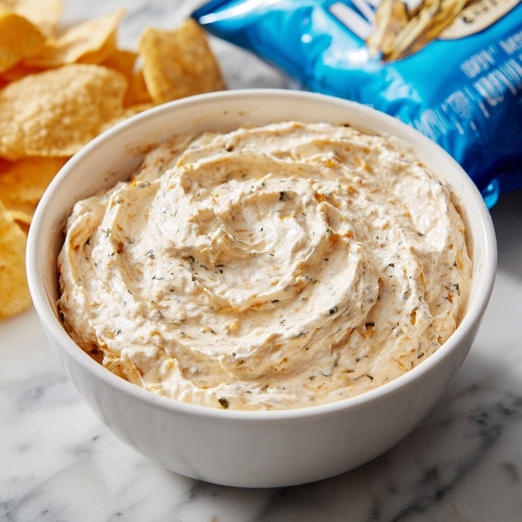 A close-up view shows a thick, creamy dip with a pale off-white color mixed with tiny bits of red and darker spices, giving a textured and slightly chunky look. The dip fills a white bowl almost to the brim, spreading smoothly but unevenly inside. A ridged light yellow potato chip is held by a woman's hand above the bowl, dipped generously and coated with the creamy mixture. The background features a blurred blue chip bag with some orange and white details, all placed on a white marbled surface. Photo taken with an iphone --ar 4:5 --v 7