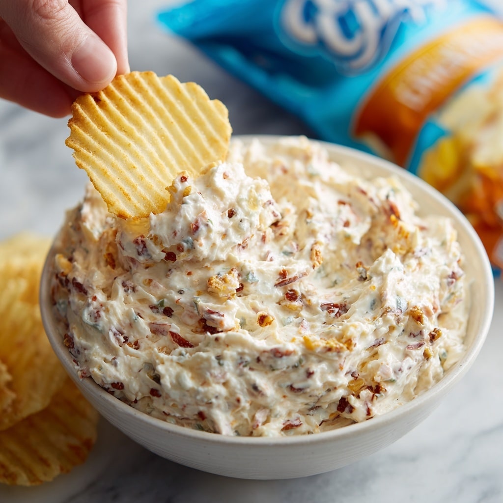 A white bowl filled with a creamy, thick dip that has a light beige color with small visible specks of herbs and bits of shredded cheese mixed throughout, showing a textured and slightly chunky surface. The dip looks rich and cheesy, with uneven swirls around the edges inside the bowl. In the background, a blue bag of party-style white corn chips is partially visible. The bowl is placed on a white marbled surface. Photo taken with an iphone --ar 4:5 --v 7