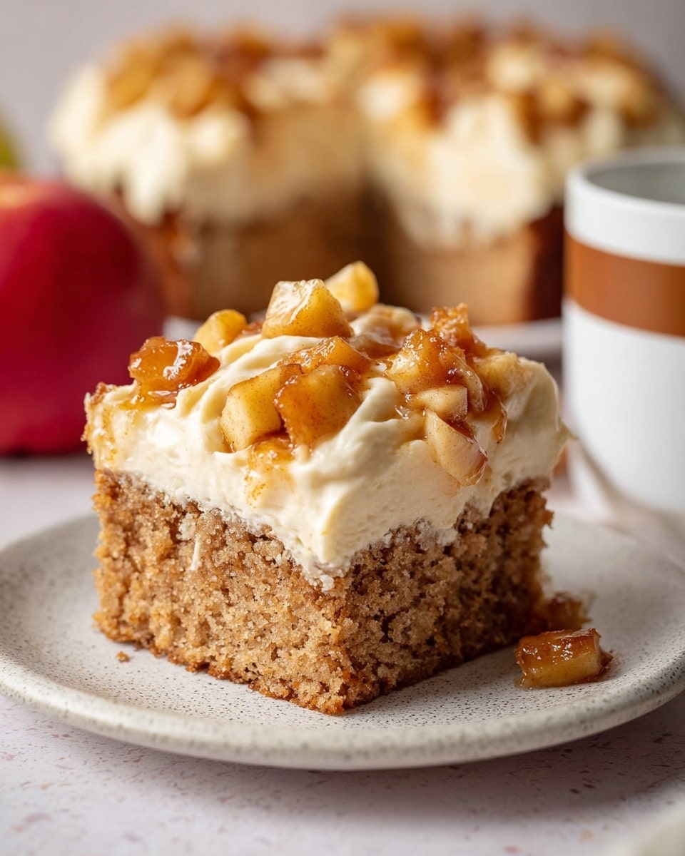 A close-up view of a single square piece of moist brown cake with a rough texture, topped by a thick layer of creamy off-white frosting with soft swirls and small, golden-brown caramelized apple chunks scattered on top. The cake is placed on a slightly textured white plate. In the blurred background, a larger frosted cake with similar apple toppings is visible, along with a red apple and a white cup with a brown stripe. The setting features a white marbled texture surface. Photo taken with an iphone --ar 4:5 --v 7