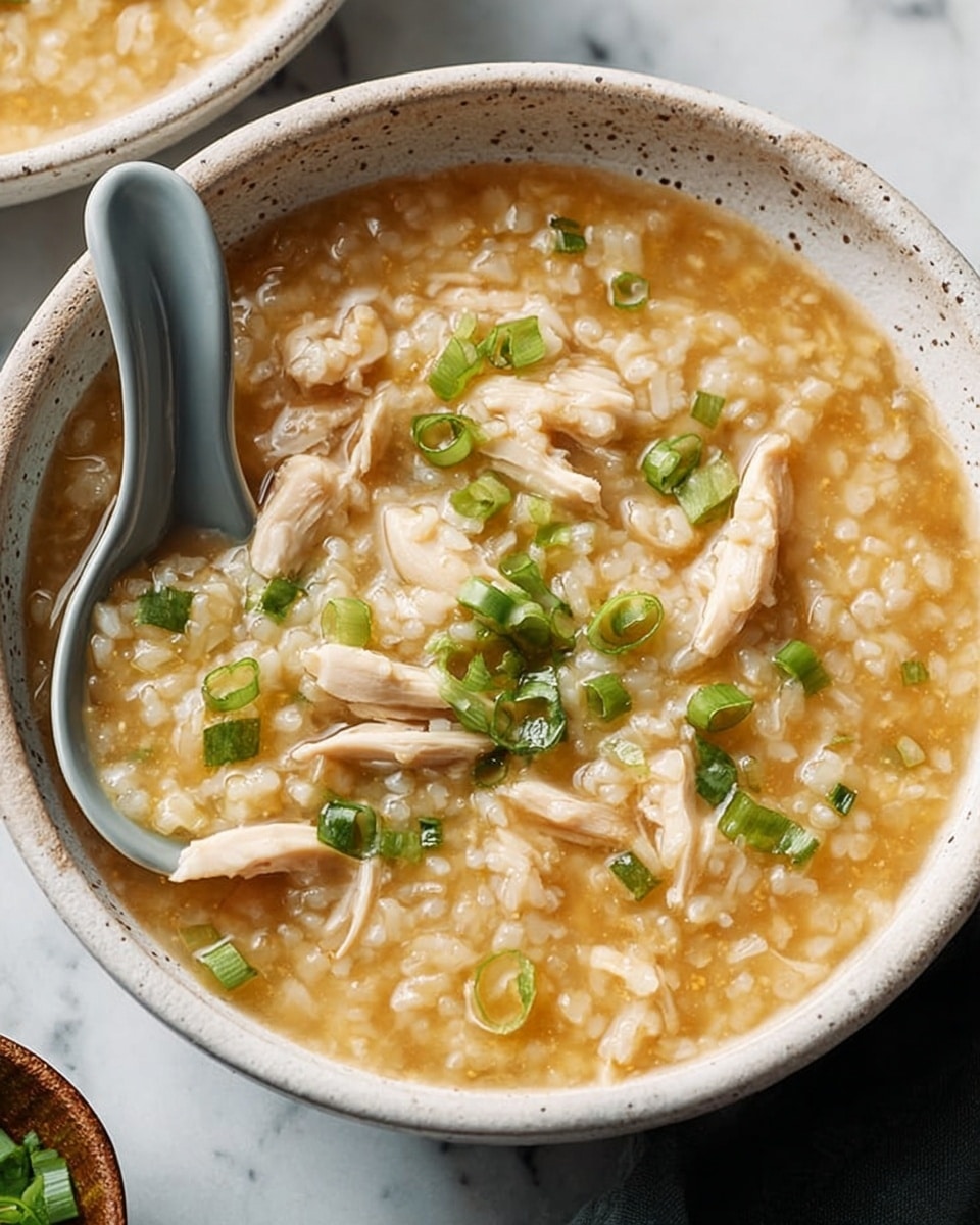 A white speckled bowl filled with a thick, light brown rice soup containing soft, light beige chicken pieces spread throughout. Small slices of green onion are sprinkled on top, adding a fresh green color contrast. A gray ceramic spoon rests inside the bowl on the left side. The bowl is placed on a white marbled surface with a wooden bowl containing coarse salt and a small wooden spoon nearby, along with some scattered green onion pieces and part of a dark blue cloth to the right. Photo taken with an iphone --ar 4:5 --v 7