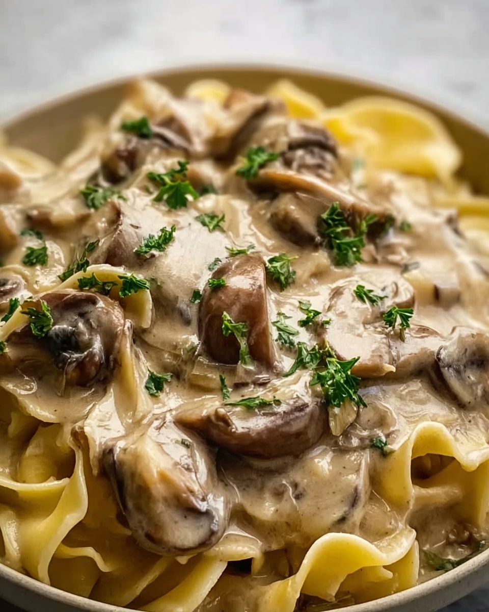 The image shows a close-up of a creamy mushroom dish with wide, flat noodles, likely pappardelle, covered in a thick, beige sauce. The sauce is rich and smooth, generously coating the noodles and mushroom slices. Small green parsley leaves are sprinkled on top, adding a fresh touch of color among the light browns and creams. The dish is served in a shallow white plate, placed on a white marbled surface. The textures of the sauce and mushrooms look soft and velvety. Photo taken with an iphone --ar 4:5 --v 7