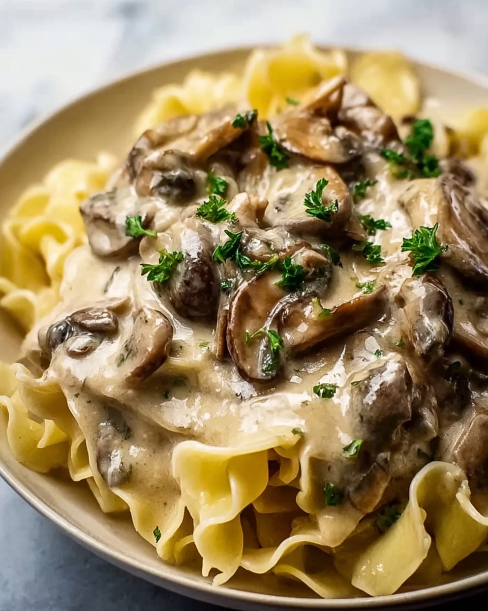 The image shows a close-up of a creamy mushroom dish with thick, wide noodles. The bottom layer is the pale yellow noodles with a soft, slightly wavy texture. On top of the noodles is a creamy beige sauce that looks thick and rich. Large pieces of brown mushrooms are mixed throughout the sauce, glossy and well coated. Small green parsley leaves are scattered on top, adding a fresh pop of color against the creamy, earthy tones. The dish is set on a white marbled surface. photo taken with an iphone --ar 4:5 --v 7