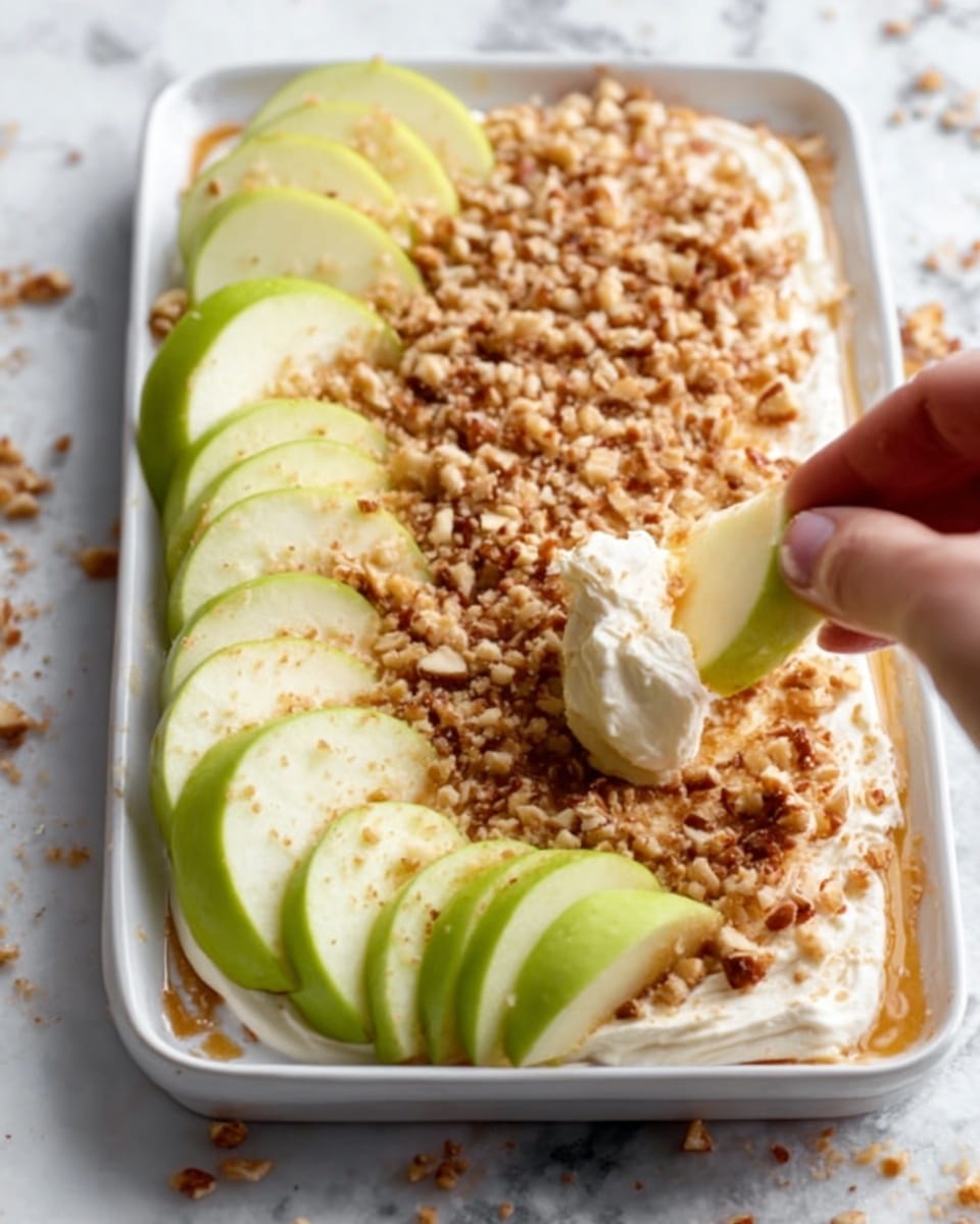 The image shows a close-up of a dish with a white rectangular plate placed on a white marbled surface. The dish contains a thick layer of smooth, creamy spread topped with a generous layer of light brown crushed nuts. Around the plate, there are fresh apple slices arranged in a neat row, with one apple slice being held by a woman's hand and dipped partly into the nut-covered spread. The apple slices are light green with shades of yellow. The overall texture highlights the crunchy nuts against the creamy spread and fresh apples. Photo taken with an iphone --ar 4:5 --v 7