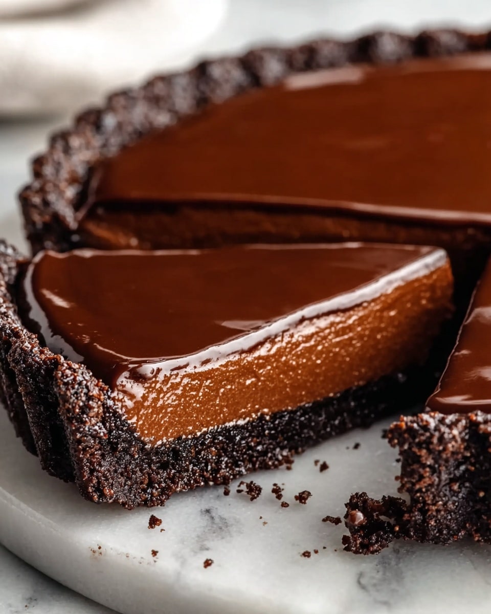 A chocolate tart with three visible layers sits on a white marbled plate. The bottom layer is a dark brown, crumbly crust that holds the tart together. The middle layer is a thick, smooth chocolate filling with a rich, creamy texture and a lighter brown color than the crust. The top layer is a shiny, glossy dark chocolate ganache that covers the filling evenly and reflects light softly. Small crumbs are scattered around the base of the tart on the plate, and pieces of chocolate lie near the plate on a wooden surface. The photo taken with an iphone --ar 4:5 --v 7