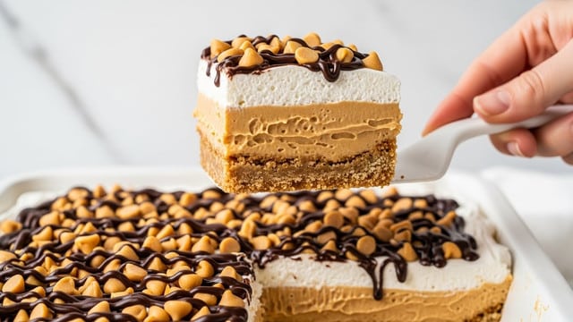 The image shows a close-up of a layered dessert in a white dish with a white marbled background. The dessert has four visible layers: at the bottom, a crumbly golden brown crust, above it a thick light brown peanut butter layer, then a fluffy white cream layer, and finally a topping of mini peanut butter chips and dark chocolate drizzle spread evenly. A woman's hand is holding a white spatula lifting one square piece from the dish, showing all the layers clearly. The scene looks clean and bright. photo taken with an iphone --ar 4:5 --v 7
