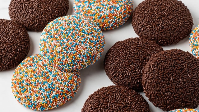 The image shows two chocolate cookie sandwiches on a white marbled surface, each coated with colorful small round sprinkles covering the top and sides. One cookie is whole while the other is broken in half, revealing a creamy white filling inside. In the background, a white mug is slightly blurred, adding depth to the image. The bright colors of the sprinkles stand out against the dark brown cookies and white filling. Photo taken with an iphone --ar 4:5 --v 7