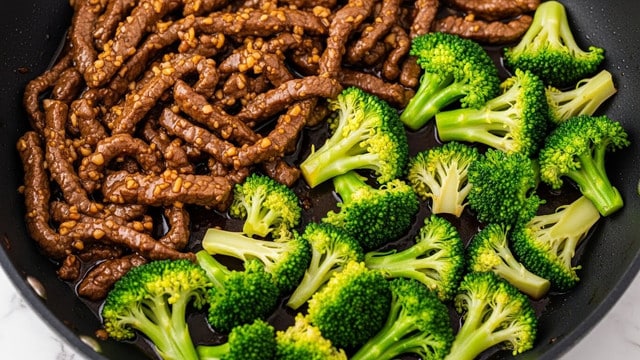 The image shows a close-up of a black pan filled with a stir-fry dish. The dish has two main layers: the first layer is tender strips of cooked brown beef with a shiny glaze from the dark sauce, and the second layer is bright green broccoli florets scattered evenly throughout the pan. The beef looks soft and juicy with small bits of garlic or ginger mixed in, while the broccoli has a fresh and slightly crisp texture. The sauce gives a glossy finish to the beef and broccoli, pooling a little bit at the bottom of the pan. The pan is placed on a white marbled surface. photo taken with an iphone --ar 4:5 --v 7