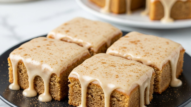 Four rectangular cake slices with a golden brown, soft crumb base are placed close together on a dark plate. Each slice is covered with a thick layer of creamy, light beige icing that smoothly drips down the sides, pooling slightly at the base. The icing has a glossy finish with tiny specks of cinnamon scattered on top, adding a touch of texture. In the blurred background, more cake pieces can be seen on a white plate, all set on a white marbled surface. Photo taken with an iphone --ar 4:5 --v 7