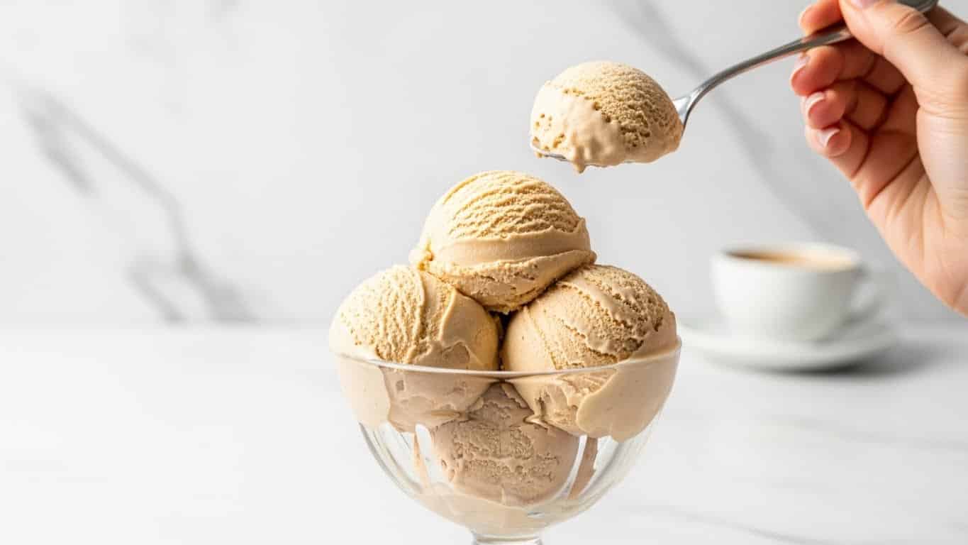 The image shows a clear glass dessert cup filled with three large scoops of creamy light brown ice cream, each scoop having a smooth and slightly textured surface. A spoon is lifting one scoop from the cup, with a woman's hand holding the spoon visible at the top right. The background is a white marbled texture, and part of a coffee cup is seen blurred in the far background. The overall scene looks bright and inviting. photo taken with an iphone --ar 4:5 --v 7