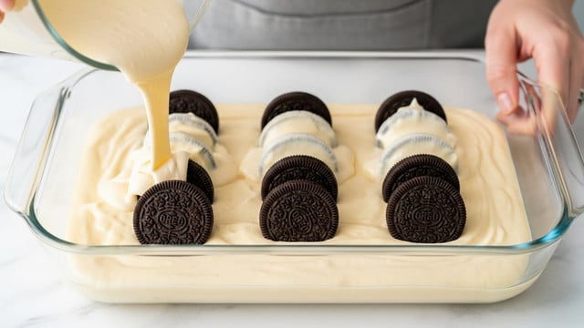 A clear rectangular glass baking dish holds three neat rows of whole sandwich cookies with dark brown outer layers and white cream filling. The cookies stand upright on a smooth, pale yellow batter that fills the bottom of the dish evenly. From above, a glass container pours a thick creamy light yellow mixture over the cookies, covering some of the cream filling and the top edges of the cookies. In the background, a woman's hand holds the glass container. The scene is set on a white marbled surface. photo taken with an iphone --ar 4:5 --v 7