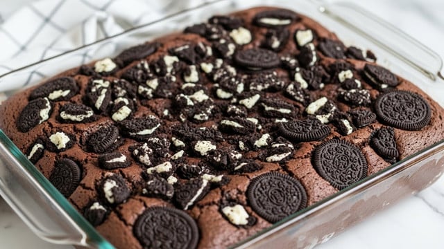 A close-up view of a thick rectangular chocolate cake baked in a clear glass pan, sitting on a white marbled surface. The cake has a deep brown color with a slightly cracked textured top, studded generously with chunks of black and white sandwich cookies, some whole and some broken, embedded on the surface. The cookies' white cream filling contrasts with the dark chocolate cake, scattered mostly on the top layer. In the background, a soft white cloth with a grey grid pattern is partly visible. photo taken with an iphone --ar 4:5 --v 7