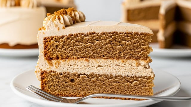 A close-up view of a two-layer slice of light brown cake with a dense, crumbly texture. The bottom layer is thick and even, topped with a middle layer of creamy, slightly darker brown frosting with a rough texture, followed by a second thick cake layer of the same light brown color and crumbly texture. The top of the slice is covered with a smooth layer of pale brown frosting, adorned with small crumbled pieces of cake or cookie on the upper left edge. This slice sits on a white plate placed on a white marbled surface with a metal fork partially visible in front. photo taken with an iphone --ar 4:5 --v 7