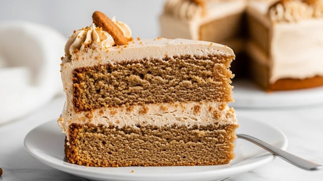 A slice of two-layer light brown cake with smooth, creamy light brown frosting covers the top and sides. Between the two moist cake layers is a thick middle layer of the same light brown frosting. The side of the slice has small crumb pieces pressed into the frosting. The cake sits on a white plate on a white marbled surface. In the background, there is a clear glass filled with milk and a silver fork lying next to the plate. photo taken with an iphone --ar 4:5 --v 7
