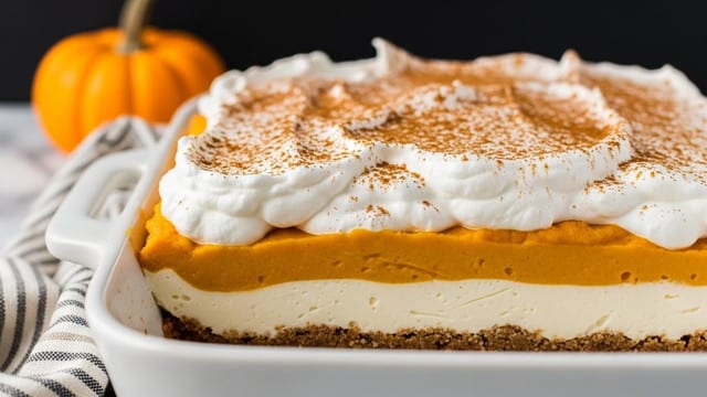 A close-up of a white baking dish filled with a layered dessert showing four distinct layers: a crumbly brown crust at the bottom, a smooth creamy white layer above it, a thick orange pumpkin puree layer in the middle, and a fluffy white whipped topping dusted with brown cinnamon powder on top. The whipped topping has soft peaks and a textured surface. The dish is placed on a surface with a white marbled texture, with a small orange pumpkin blurred in the dark background. In the foreground, a striped cloth is partially visible. Photo taken with an iphone --ar 4:5 --v 7